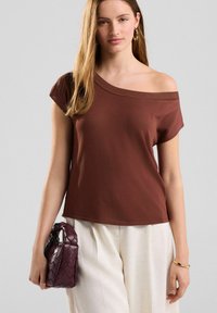T-shirt marron à épaules dénudées en tissu doux, associé à un pantalon large crème. Porte un petit sac à main tressé bordeaux.