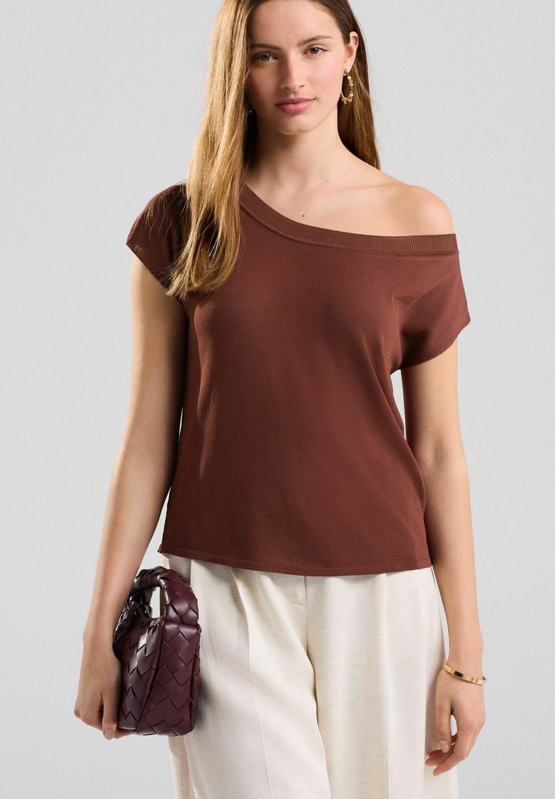 T-shirt marron à épaules dénudées en tissu doux, associé à un pantalon large crème. Porte un petit sac à main tressé bordeaux.