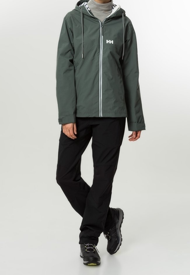 Veste imperméable vert foncé avec une fermeture éclair sur le devant, capuche ajustable et logo blanc. Associée à un pantalon noir et des chaussures noires robustes.