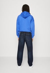 Sudadera azul con cordón, corte holgado; pantalones de mezclilla oscuros con bolsillos traseros; modelo vistiendo zapatillas blancas.