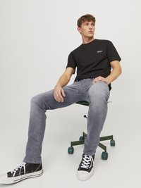Schwarzes Kurzarm-T-Shirt mit weißem Text, kombiniert mit hellgrauen Slim-Fit-Jeans und schwarzen High-Top-Sneakern. Vor einem schlichten Hintergrund.