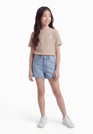 Mlada djevojka s dugom tamnom kosom, odjevena u bež majicu, svijetloplave kratke traperice i bijele tenisice, stoji i smiješi se gledajući u stranu.