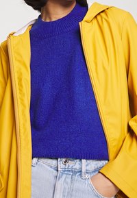 Gul vattentät jacka med huva, bärs över en strukturerad blå tröja. Har en ribbad krage och ljusblå denimjeans.