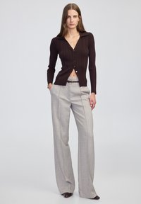 Eine Frau steht mit den Händen in den Taschen und trägt eine dunkelbraune gerippte Strickjacke und hochgeschnittene hellgraue weite Hosen.