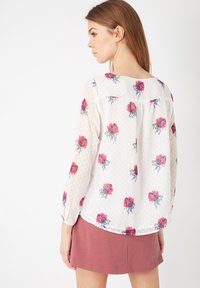 Blusa blanca translúcida con patrón floral rojo, textura de puntos, escote redondeado y mangas largas, combinada con una falda rosa.