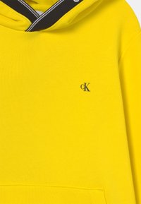 Felpa gialla con cappuccio in cotone, caratterizzata da un collo a righe bianche e nere e un piccolo logo blu "CK" sul petto.