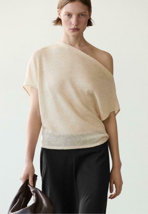 Frau trägt ein beiges Ein-Schulter-Top und schwarze Hose, hält eine braune Lederhandtasche in der linken Hand.