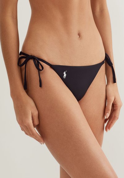 Μαύρο κάτω μέρος μπικίνι με δεσίματα στο πλάι και λευκό λογότυπο παίκτη πόλο, φορεμένο στους γοφούς μιας γυναίκας σε ουδέτερο φόντο.