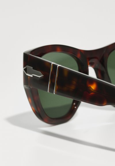 Óculos de sol tartaruga com lentes verde-escuras, apresentando uma armação espessa e um detalhe prateado na haste. Superfície suave e design angular.