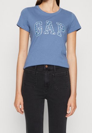 Kobieta w niebieskiej koszulce z krótkim rękawem z napisem "GAP" w białe kwiatowe litery oraz w czarnych, wysokich spodniach dżinsowych z naszywanymi kieszeniami z przodu.