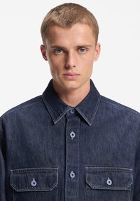 Camisa de mezclilla oscura con cuello clásico, dos bolsillos frontales con cierres de botones y costuras en contraste por toda la prenda.