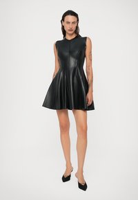 Γυναίκα με τατουάζ φορώντας ένα μίνι φόρεμα από μαύρο δερμάτινο υλικό χωρίς μανίκια και μαύρες μυτερές γόβες, στέκοντας μπροστά σε λευκό φόντο.