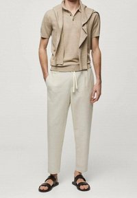 Homme portant un pull à manches courtes beige avec un gilet assorti, un pantalon à cordon de serrage beige clair et des sandales noires, se tenant les mains dans les poches.