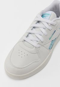 Bijele kožne tenisice s teksturiranim plavim naglaskom, ravnim vezicama, perforiranim prstima i suptilnim Reebok logotipom sa strane.