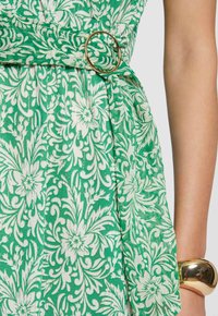 Grünes Kleid mit floralem Muster aus strukturiertem Stoff, mit einem goldenen, runden Gürtel und einer fließenden Silhouette.