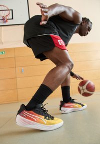 Basketballsko med en sort overdel, gul-orange gradientdesign, hvid sål og sorte snørebånd. Har tekstureret materiale og slankt design.