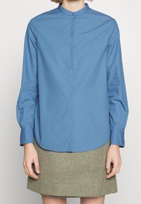 Blå långärmad skjorta med ståkrage, framknäppning och rundad nederkant, tillsammans med en grå kjol. Slät tygtextur.