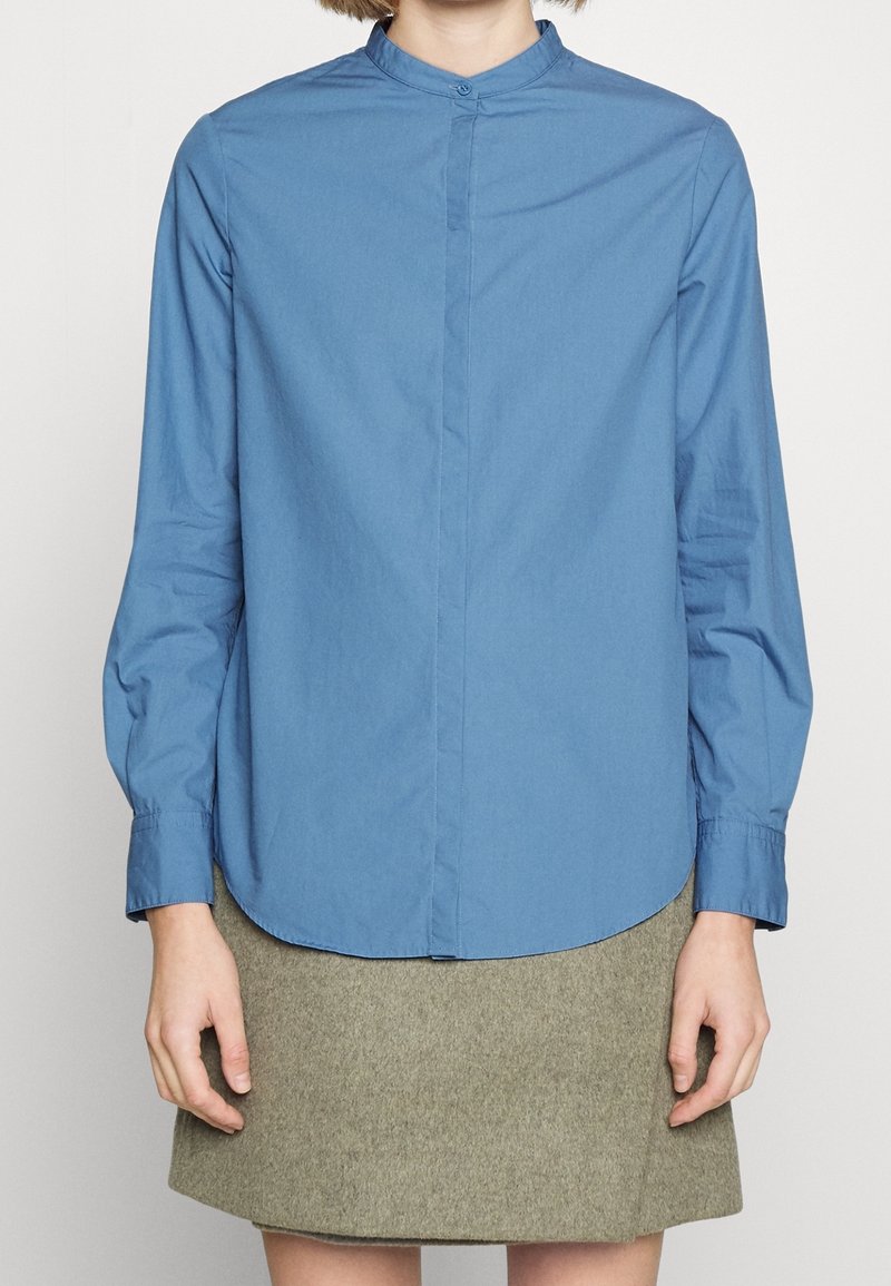 Blå långärmad skjorta med ståkrage, framknäppning och rundad nederkant, tillsammans med en grå kjol. Slät tygtextur.