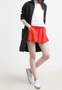 Rote Shorts mit einem fließenden Design, kombiniert mit einer weißen Bluse aus Spitzenstoff und einem langen schwarzen Mantel. Weiße Schnürschuhe vervollständigen das Outfit.