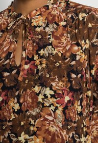 Chemisier floral sur fond sombre, arborant de grandes fleurs brunes, oranges et jaunes, avec un décolleté en trou de serrure et un détail à nouer. Tissu doux et texturé.