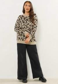 Beżowy sweter z wzorem w panterkę, z prążkowanym dołem i mankietami, w połączeniu z szerokimi czarnymi spodniami i masywnymi czarnymi butami, stojący w pobliżu neutralnej ściany.