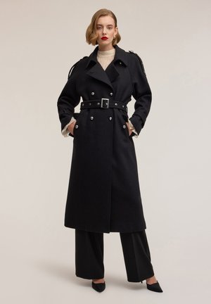 Zwarte wollen trenchcoat met dubbelgeknoopte sluiting, tailleband, zilveren knoopaccenten en grote zakken. Model draagt zwarte broek en hakken.