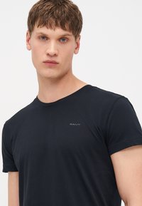 Joven con cabello castaño corto que lleva una camiseta negra lisa de GANT mirando hacia adelante con fondo blanco.