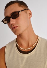 Νεαρός άνδρας με κοντά μαλλιά, φοράει σκούρα ορθογώνια γυαλιά ηλίου, αμάνικο μπεζ τοπ πάνω από ένα σκουρόχρωμο εσώρουχο και περιδέραιο με χάντρες, με φόντο φωτεινό.