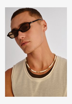 Noor mees lühikeste juustega, kellel on selged tumedad ristkülikukujulised päikeseprillid, beež värvi varrukateta top tumeda särgi peal ning helmestega kaelakee heleda tausta ees.