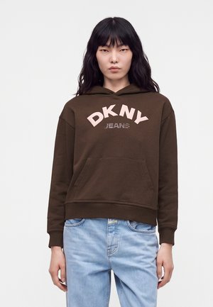 Personne portant un sweat à capuche marron foncé avec le texte "DKNY JEANS" en rose clair et un jean bleu clair sur un fond blanc uni.