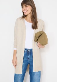 Femme souriante tenant une veste vert olive pliée, portant un cardigan beige par-dessus une chemise blanche et un jean bleu, sur fond blanc.