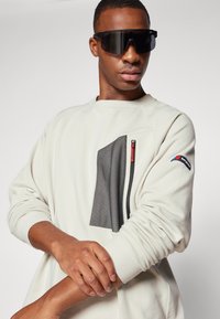 Light cream sweatshirt with textured black mesh pocket, red zipper detail, and ribbed cuffs. Worn with oversized black sunglasses.