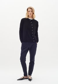 Černý pletený cardigan s knoflíkovými detaily, kombinovaný s úzkými námořnickými kalhotami a černými špicatými balerínami. Modelka stojí s rukama v kapsách.