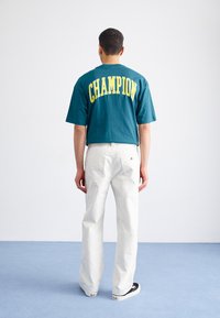 Camiseta de algodón color verde azulado con letras amarillas "CHAMPION" en la parte de atrás, combinada con pantalones rectos blancos y zapatillas negras.