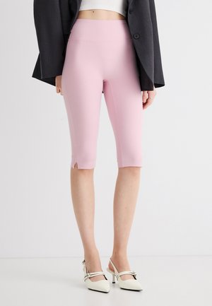 Persoană care poartă pantaloni scurți roz, mulați, până la genunchi, top alb scurt, sacou negru și pantofi albi cu toc și barete în spate, pe un fundal simplu.