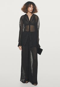 Completo nero trasparente composto da una lunga camicia con bottoni e pantaloni a gamba larga con texture plissettata. Il tessuto è leggero e traslucido.