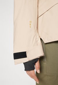 Beige vanntett jakke med svart borrelås på ermet, med gull A.W.S. Pro-logo. Glatt tekstur, skreddersydd passform og slank design.