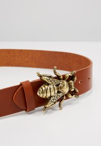 Brown leather belt featuring a large, detailed, golden bee buckle. Smooth texture with visible stitching and adjustable length.