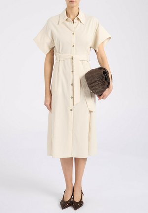 Donna che indossa un vestito midi crema con bottoni e cintura, tiene una clutch marrone strutturata e indossa tacchi a punta marrone scuro.