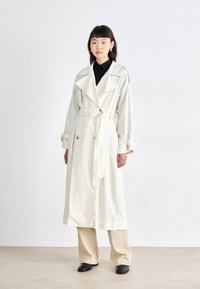 Manteau trench crème avec un design à double boutonnage, large col et ceinture. Présente des manches longues, des accents de boutons et une coupe décontractée. Chemise noire en dessous.