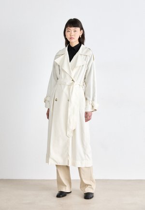 Cremfarbener Trenchcoat mit doppelreihigem Design, breitem Kragen und Gürtel. Hat lange Ärmel, Knopfdetails und eine lockere Passform. Schwarzfarbenes Hemd darunter.