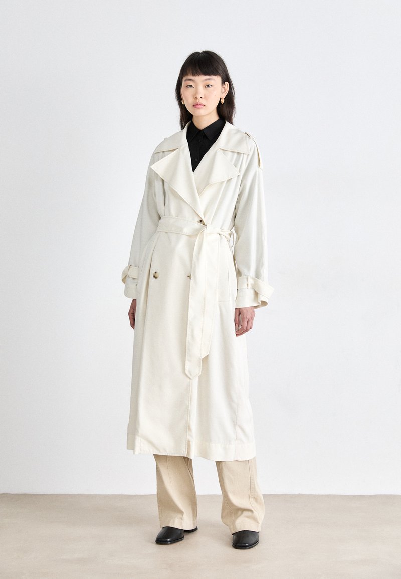 Manteau trench crème avec un design à double boutonnage, large col et ceinture. Présente des manches longues, des accents de boutons et une coupe décontractée. Chemise noire en dessous.