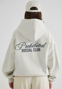 Hoodie oversized grigio chiaro con cappuccio, con la scritta "Prohibited Social Club" ricamata in blu scuro sul retro. Tessuto morbido.