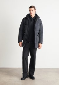 Black puffer jacket with a hood, featuring a zip and snap closure. Worn over a black polo shirt and dark trousers, paired with black boots.
