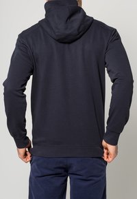 Námořnická modrá mikina s kapucí, dlouhými rukávy a žebrovaným límcem. Hladká textura, bez viditelných vzorů nebo kovových detailů.