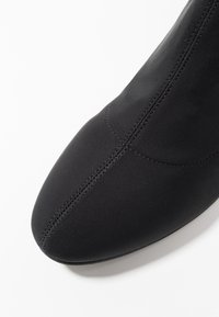 Botín negro con un diseño elegante, hecho de tela elástica. Presenta una punta afilada y detalles de costura minimalistas. Textura suave.