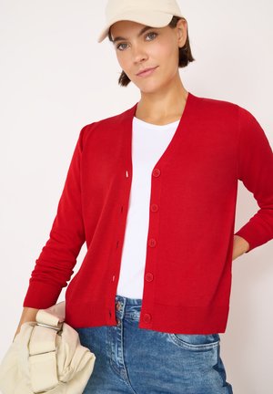 Rode cardigan met V-hals, knopen aan de voorkant en lange mouwen. Zacht breimateriaal, gecombineerd met een wit T-shirt en blauwe denim shorts.