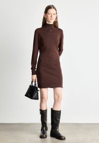 Vestido de punto marrón de manga larga con cuello alto, combinado con botas altas marrones y un pequeño bolso negro. Textura suave, forma ajustada.