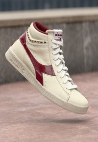 High-top sneakers in cream leather with maroon accents. Features a round toe, white laces, and textured sole. Decorative gold studs included.