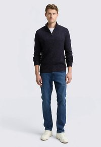 Marineblauwe gebreide trui met halve rits, lange mouwen, gestructureerd patroon, gedragen over een wit overhemd, gecombineerd met een blauwe spijkerbroek en witte sneakers.